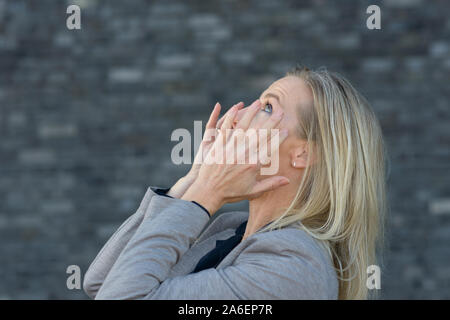 Femme blonde hurlant avec les mains sur ses joues vu de son côté en close-up, debout à l'extérieur en manteau gris à l'arrière-plan gris foncé Banque D'Images