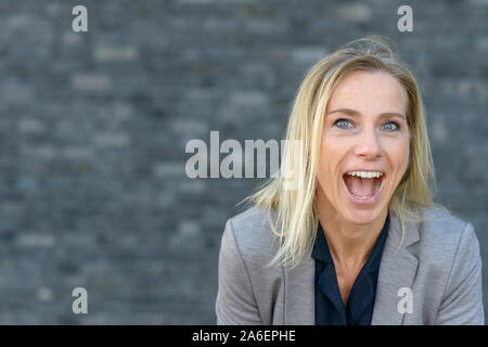 Vive heureux belle blue-eyed woman cheering comme elle rit à l'appareil photo à l'extérieur contre un mur avec copie espace Banque D'Images