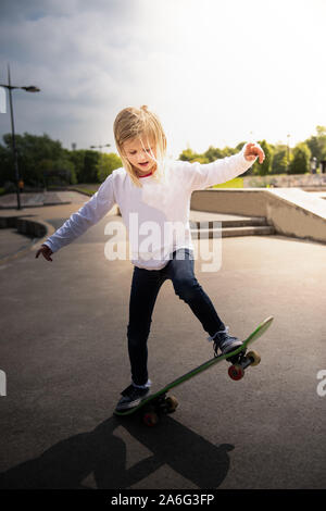 Une jolie petite fille aux cheveux blonds à l'échelle locale et d'un planchodrome pratique équestre autour sur son skateboard tout en écoutant de la musique Banque D'Images