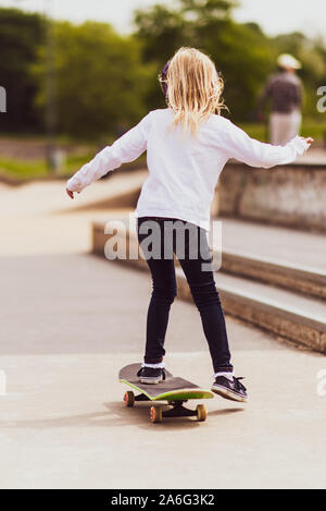 Une jolie petite fille aux cheveux blonds à l'échelle locale et d'un planchodrome pratique équestre autour sur son skateboard tout en écoutant de la musique Banque D'Images