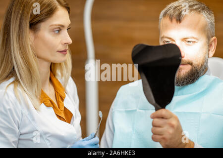 L'homme à la recherche sur le miroir dentaire, satisfait de ses dents, lors d'une consultation médicale avec femme dentiste au cabinet dentaire Banque D'Images