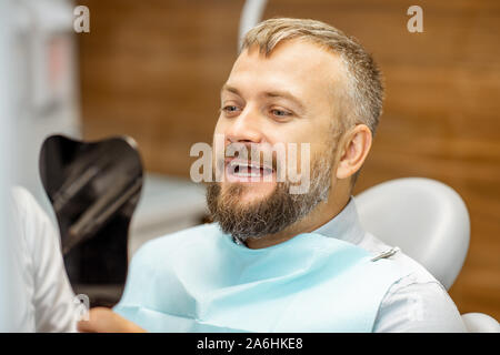 L'homme à la recherche sur le miroir dentaire, satisfait de ses dents, lors d'une consultation médicale au cabinet dentaire Banque D'Images