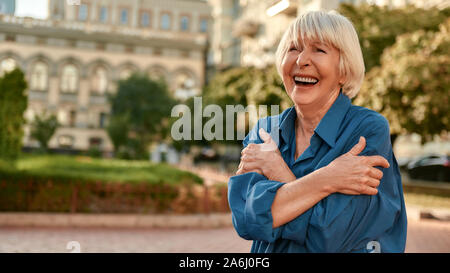 Je suis optimiste. Belle senior woman in casual wear serrant ses épaules et sourit tout en se tenant à l'extérieur sur une journée ensoleillée Banque D'Images