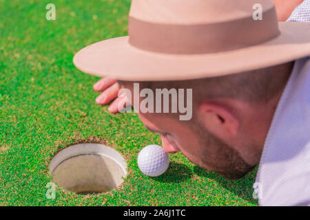 Un jeune homme avec une barbe et un chapeau, en étroite collaboration l'observation d'une balle de golf sur un terrain vert. Concept d'une journée de détente et de loisirs. Vue d'en haut Banque D'Images