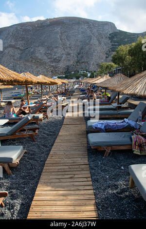 Kamari, Grèce - 14 juillet 2019 : un conseil de marche, à travers le sable noir entre les parasols et transats sur la plage de Kamari Banque D'Images