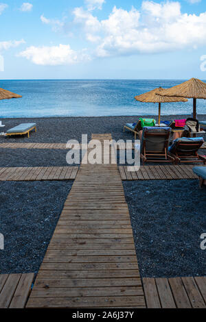 Kamari, Grèce - 14 juillet 2019 : un conseil de marche, à travers le sable noir entre les parasols et transats sur la plage de Kamari Banque D'Images