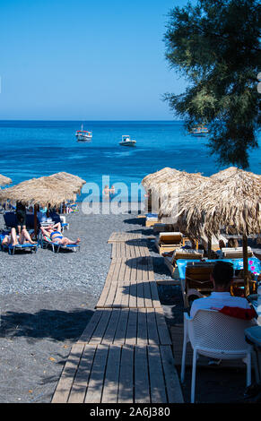 Kamari, Grèce - 14 juillet 2019 : un conseil de marche, à travers le sable noir entre les parasols et transats sur la plage de Kamari Banque D'Images