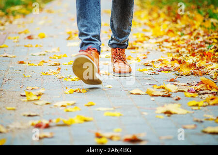 Les jambes d'un homme en bottes marron en marchant le long du trottoir jonché de feuilles mortes. La notion de chiffre d'affaires de la saison de l'année. Background météo Banque D'Images