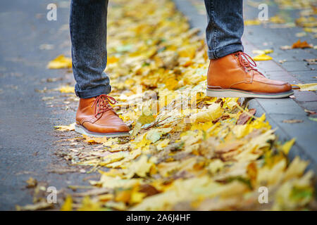 Les jambes d'un homme en bottes marron en marchant le long du trottoir jonché de feuilles mortes. La notion de chiffre d'affaires de la saison de l'année. Background météo Banque D'Images