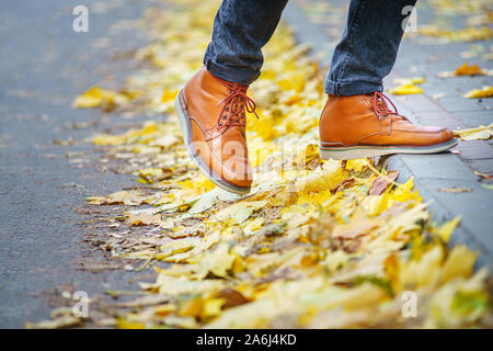 Les jambes d'un homme en bottes marron en marchant le long du trottoir jonché de feuilles mortes. La notion de chiffre d'affaires de la saison de l'année. Background météo Banque D'Images