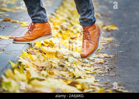 Les jambes d'un homme en bottes marron en marchant le long du trottoir jonché de feuilles mortes. La notion de chiffre d'affaires de la saison de l'année. Background météo Banque D'Images