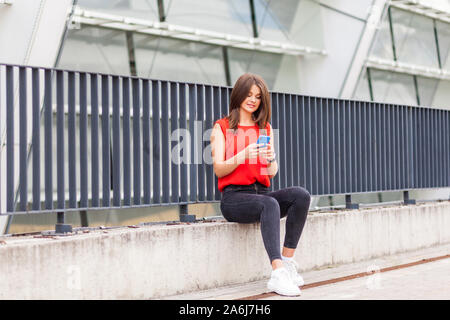 Internet mobile dans des lieux publics. Portrait of positive charming brunette woman in stylish chemise rouge assis près de clôture métallique, les sms sur téléphone ou scro Banque D'Images