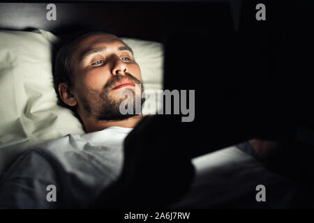 Beau jeune homme fatigué et avec une barbe ne peut pas dormir et regarde quelque chose sur sa tablette de nuit. Banque D'Images