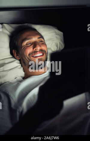 Handsome smiling caucasian male barbu au lit la nuit tout en regardant quelque chose sur sa tablette. Banque D'Images