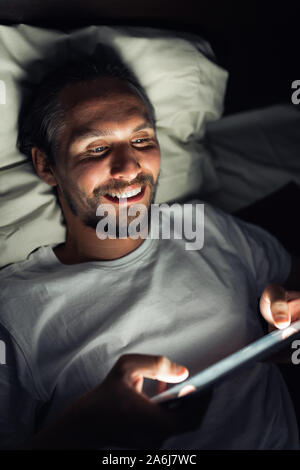 Handsome smiling caucasian male barbu au lit la nuit tout en regardant quelque chose sur sa tablette. Banque D'Images