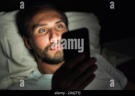 Beau jeune homme fatigué et avec une barbe ne peut pas dormir et regarde quelque chose sur son téléphone dans la nuit. Banque D'Images