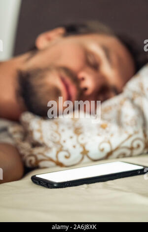 Photo en gros plan d'un jeune homme de dormir dans le lit. Téléphone mobile avec un écran clignotant blanc est visible au premier plan. Banque D'Images