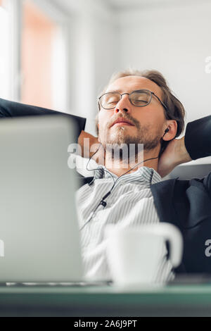 Jeune homme essaie de se détendre et de dormir tout en écoutant de la musique au travail. Tasse à café et un ordinateur visible au premier plan. Banque D'Images