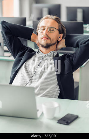 Jeune homme essaie de se détendre et de dormir tout en écoutant de la musique au travail. Tasse à café, ordinateur et un téléphone mobile visible dans l'avant-plan. Banque D'Images