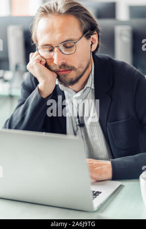 Les jeunes fatigué et ennuyé bel homme essaie de se détendre et de dormir au travail. Il porte un casque noir. Banque D'Images