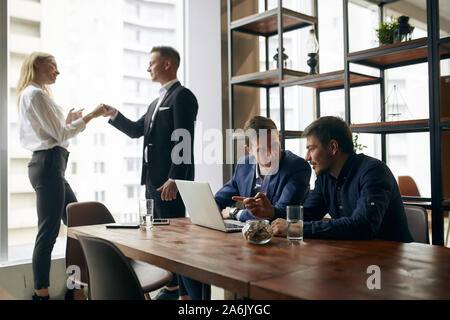Beaux hommes assis au bureau travailler sur ordinateur portable, tandis que l'homme gai et belle blonde woman shaking hands, flirter dans l'arrière-plan de la pho Banque D'Images
