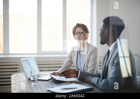 Portrait de deux jeunes entrepreneurs de discuter affaires projet tandis qu'assis à table de réunion derrière mur de verre, se concentrer sur smiling woman looking at pa Banque D'Images