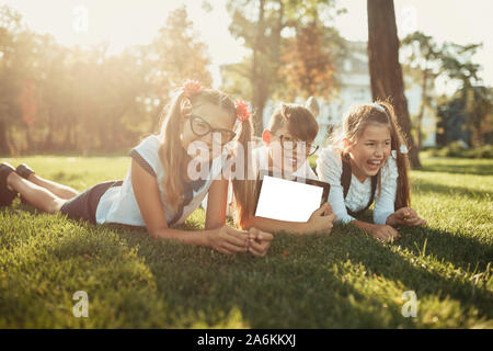 Trois amis d'âge souriants sont allongés sur l'herbe et regardant la caméra. Dans les mains d'enfants une tablette Banque D'Images