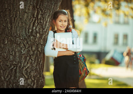 Une belle fille d'âge scolaire se tient à un arbre et tenant une tablette. Habillée d'une robe d'écolière Banque D'Images