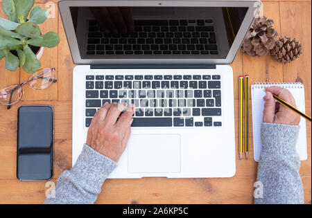Voir ci-dessus de senior woman's hands working on laptop et l'écriture. Table en bois. Bureau extérieur de rechange. Téléphone cellulaire et les lunettes près de sa Banque D'Images
