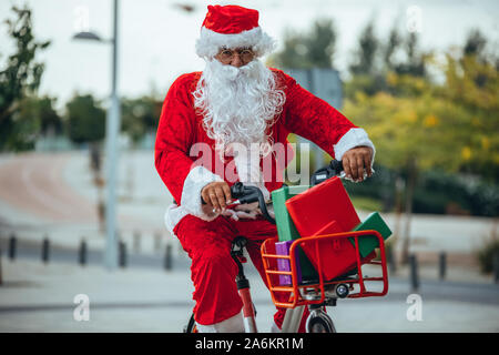 Stock photo du Père Noël avec des cadeaux dans le vélo et sa valise rouge sur son dos. Temps de Noël Banque D'Images