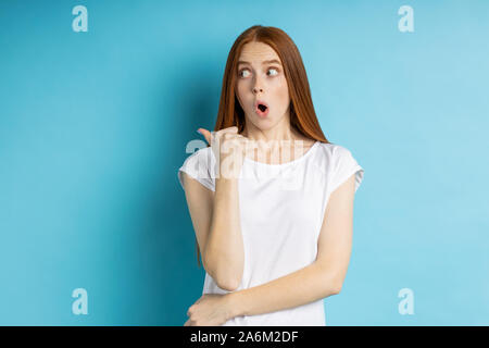 Surpris et choqué jeune rousse modèle féminin en t-shirt blanc à côté d'écoute avec les yeux, bouche ouverte en montrant l'espace de copie pour votre texte ou adve Banque D'Images