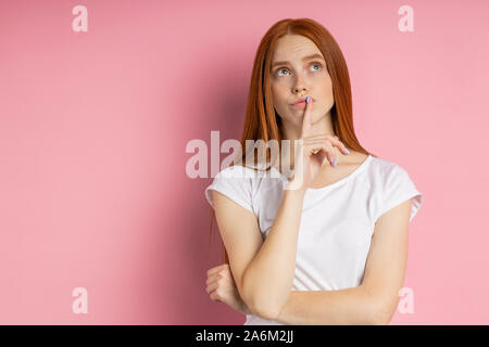 Pensive young caucasian ginger femelle avec des taches de rousseur peau cheveux longs portant des T-shirt blanc à la recherche jusqu'à l'expression perplexe, touchant ses lèvres de forefi Banque D'Images