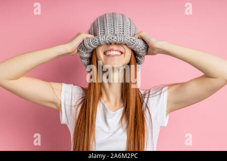 Close up portrait of cheerful ginger girl with long hair s'amusant, couvrant les yeux avec grande boucle Chapeau tricoté, tout sourire avec des dents blanches isoler Banque D'Images