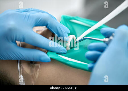 Fille charmante dans des lunettes et un enrouleur joue avec un barrage en caoutchouc vert dans une clinique dentaire. Dentiste en bleu gants en latex est nettoyer son tee Banque D'Images