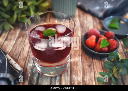 Verre rouge transparent en verre avec des glaçons en forme de coeur, fraises et autre décoration en arrière-plan on wooden table Banque D'Images