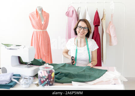 Jeune fille slim designer couturière portant des lunettes est assis à son bureau avec une machine à coudre fil et coupe un morceau de tissu pour coudre les nouveaux produits Banque D'Images