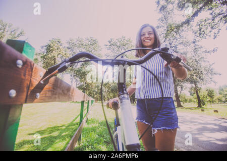 Portrait of attractive young woman in shorts jeans avec vélo dans le parc le jour d'été. Low angle view. Banque D'Images