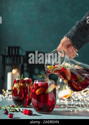 Hiver main de femme verse dans des verres avec des fruits de la sangria, canneberges et romarin coupe. Vacances de Noël avec l'arrière-plan foncé bougie, guirlande de décoration. L'espace pour copier du texte ou dessin. La verticale Banque D'Images