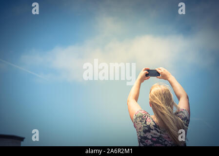 Young Caucasian lady taking photo à l'aide de son téléphone intelligent contre le ciel bleu Banque D'Images