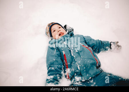 Happy boy sticking out tongue en position couchée dans la neige en hiver Banque D'Images