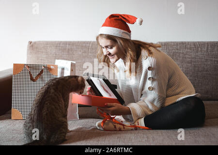 Happy girl in santa hat opening christmas gift box et jouer avec chat. Jeune femme tenant présent et montrer à son chat Banque D'Images