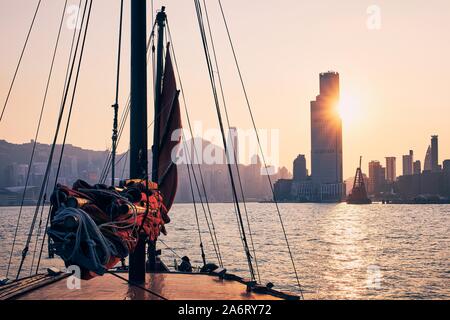 Traversée en bateau jonque traditionnelle contre Victoria Harbour Hong Kong cityscape avec gratte-ciel au coucher du soleil doré. Banque D'Images