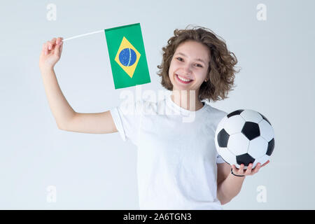 Jeune femme heureuse avec un ballon de football dans ses mains et le drapeau du Brésil debout sur un fond gris. Fan de football. Banque D'Images