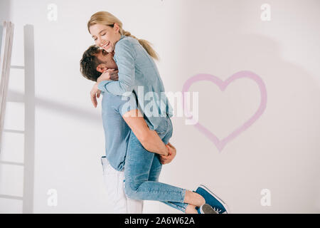 Happy young man holding petite amie sur les mains près du coeur rose dessiné sur mur blanc Banque D'Images