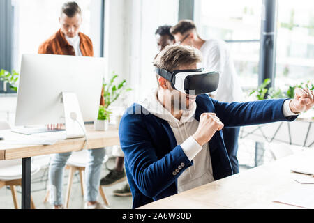 Jeune homme d'imitation de lutte lors de l'utilisation de casque vr, et multiculturelle des collègues qui travaillent dans office Banque D'Images