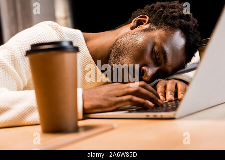 Focus sélectif de épuisé african american businessman sleeping sur le lieu de travail près de l'ordinateur portable et gobelet jetable de nuit in office Banque D'Images