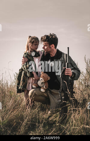 Handsome man holding gun près de l'enfant avec ours en peluche dans le champ, concept post apocalyptique Banque D'Images