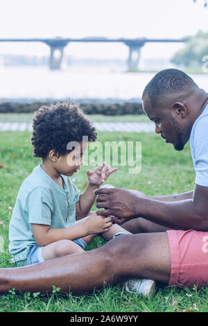 Young african american montrant fils assis sur balle de baseball en pelouse park Banque D'Images