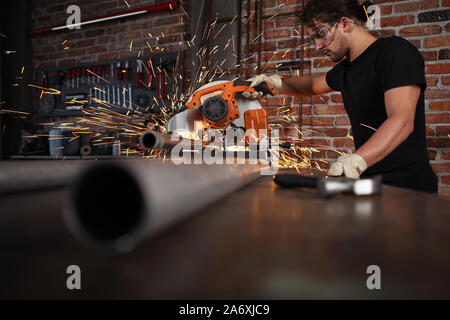 L'homme travail dans l'atelier de maison avec garage à onglets, lunettes et gants de construction, scier le métal pipe fait des étincelles, bricolage et artisanat concept Banque D'Images