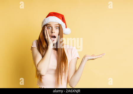 Stupéfait caucasian ginger girl in Santa hat et crème couleur t shirt, holding copy space, posant avec open palm, de montrer ou de présenter votre produit, ite Banque D'Images